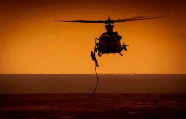 A man rappelling from a helicopter, with both silhouetted against an orange sky, and the brown dirt beneath them